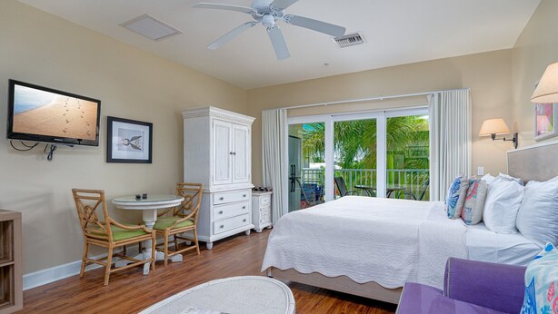 Um quarto com cama, uma cadeira, cômoda, televisão e ventilador de teto.