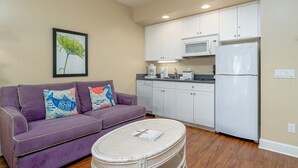 Um espaço compacto para viver, com sofá roxo, mesa de centro de vime, cozinha compacta com armários brancos, micro-ondas e geladeira.