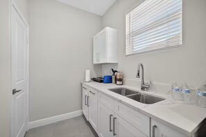 Una cocina con mostrador blanco, un lavamanos, una ventana con persianas y una puerta.