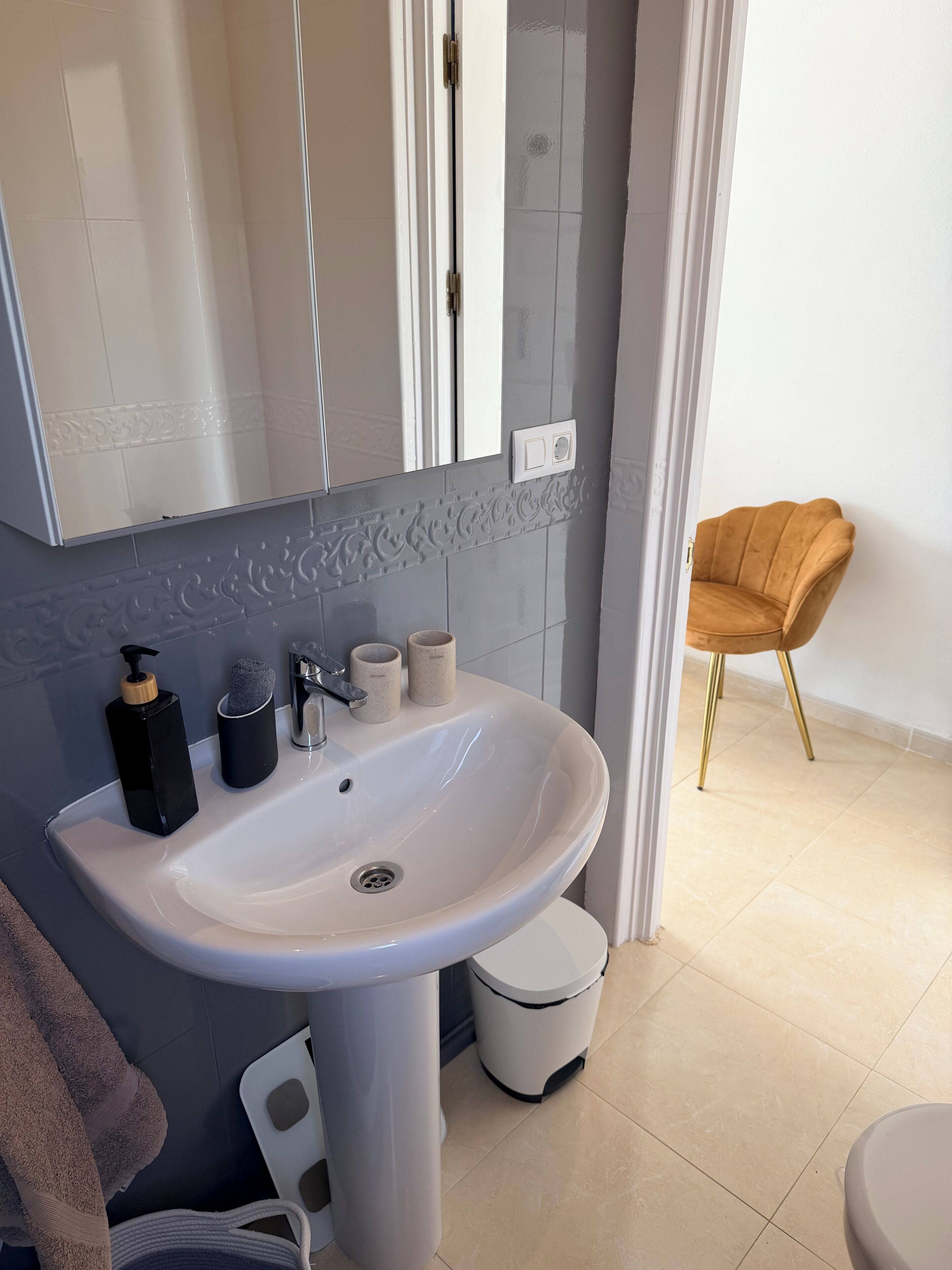 Un bagno con un lavandino bianco, uno specchio, un asciugamano e un cestino.