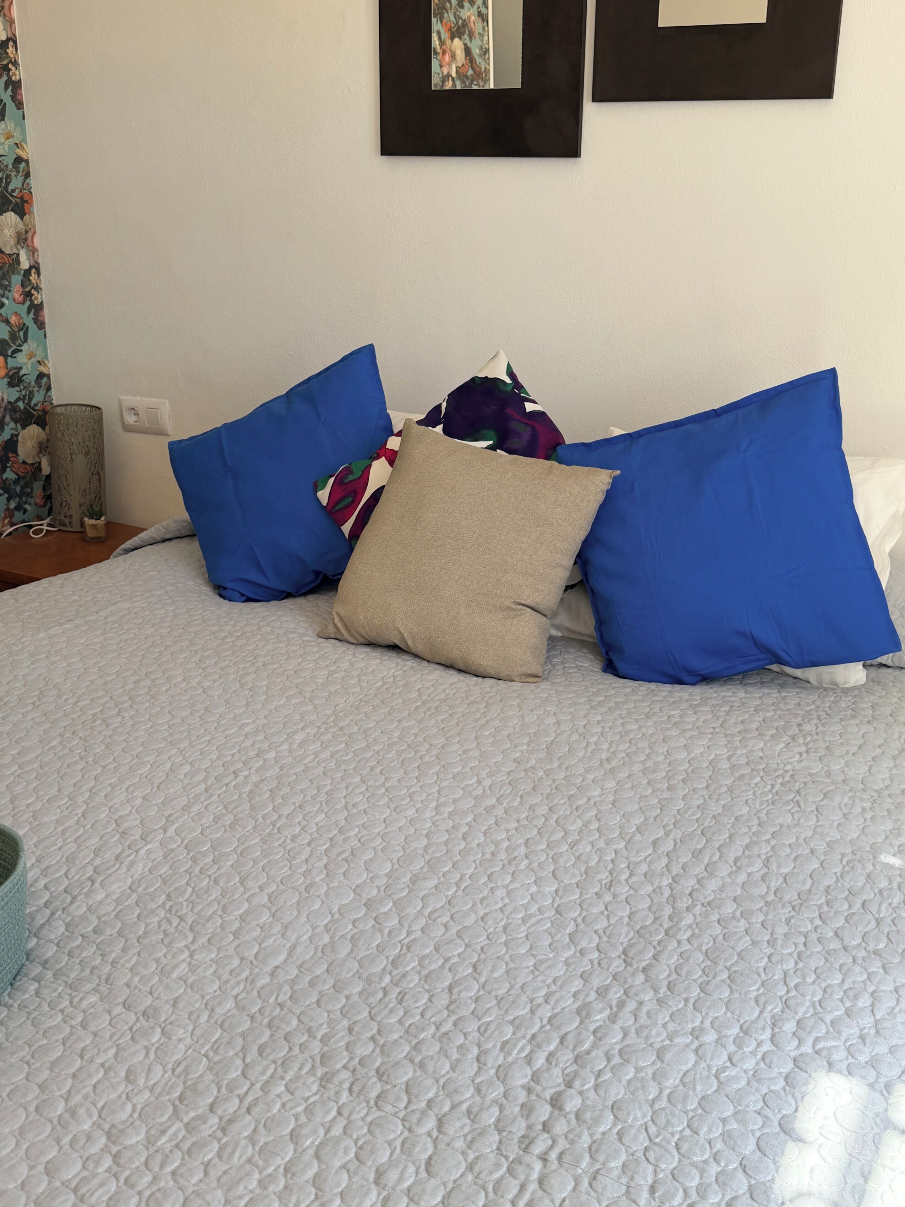 A bed with a textured white cover, blue and beige pillows, and a small decorative item on the side.