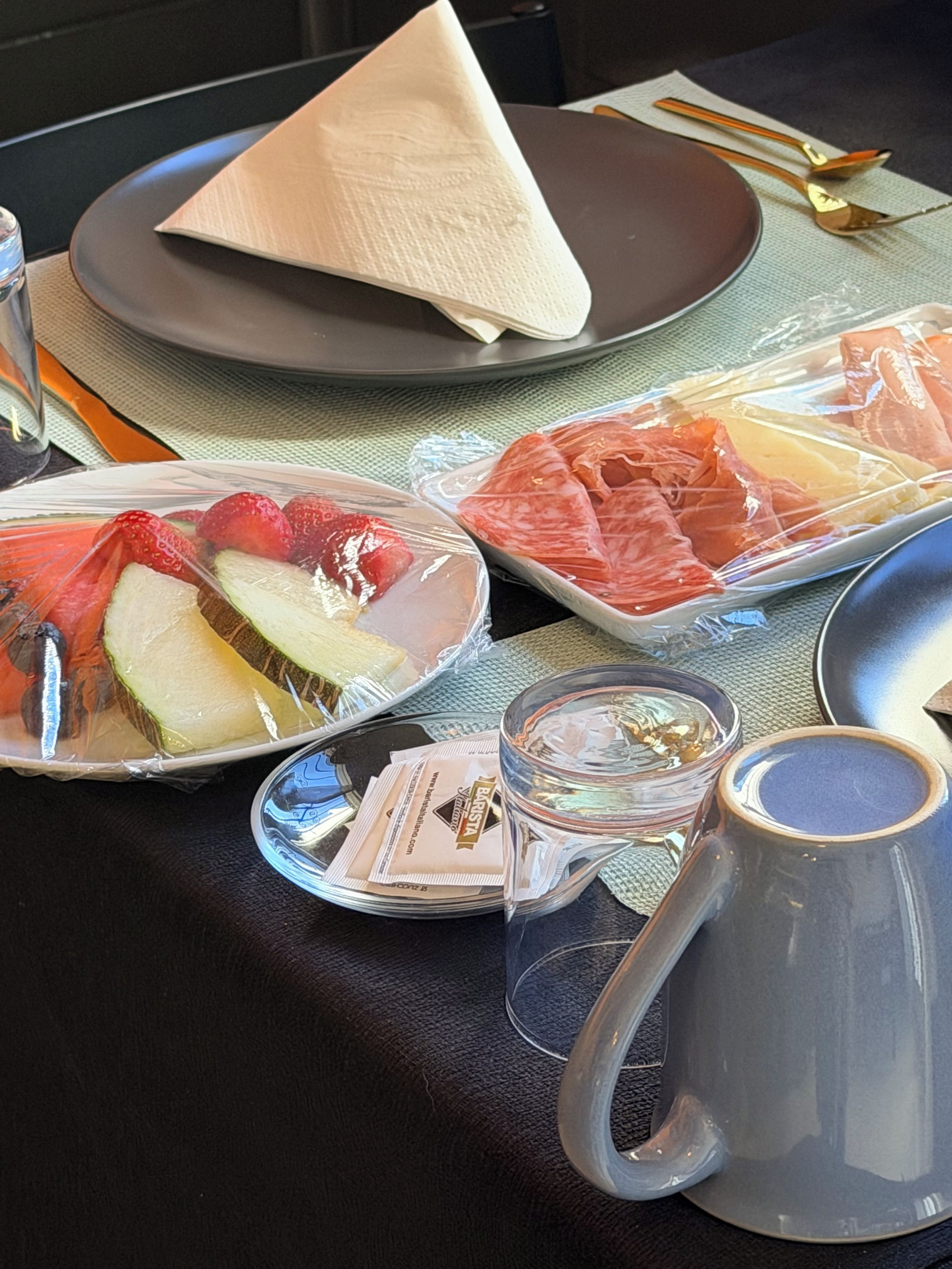 Une table avec une assiette de fruits coupés, un plat de viande emballé, un verre et une serviette sur une nappe sombre.
