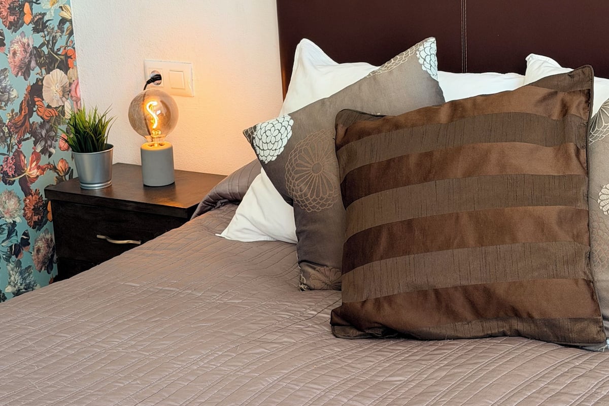 A neatly made bed with a striped pillow, a bedside lamp, and a floral-patterned curtain.