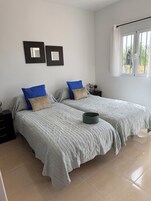 A bedroom with two beds, a window with curtains, and a small table with a lamp.