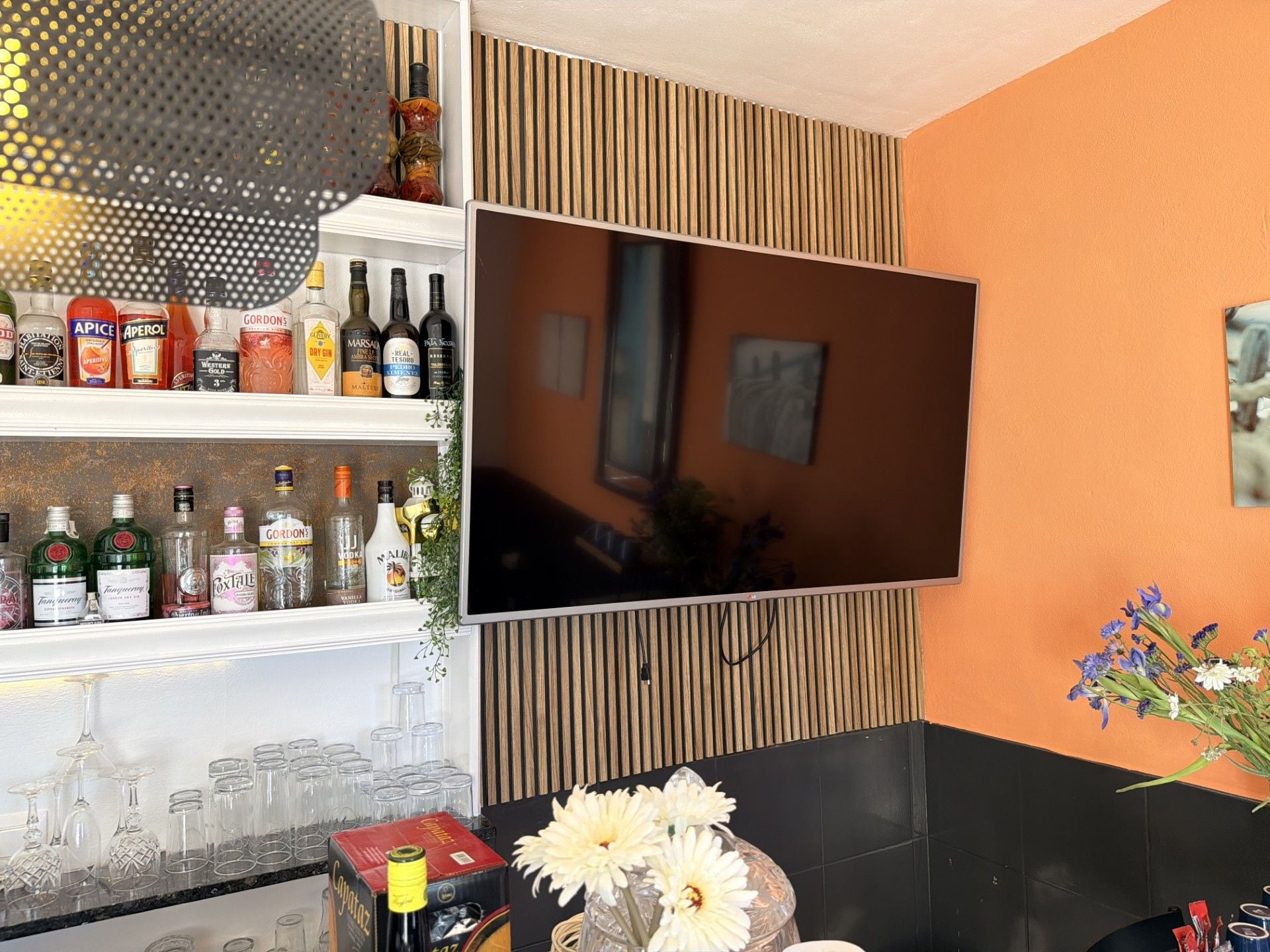 Una zona de bar con un televisor montado en la pared, estantes con diversas botellas de licor y un jarrón con flores.
