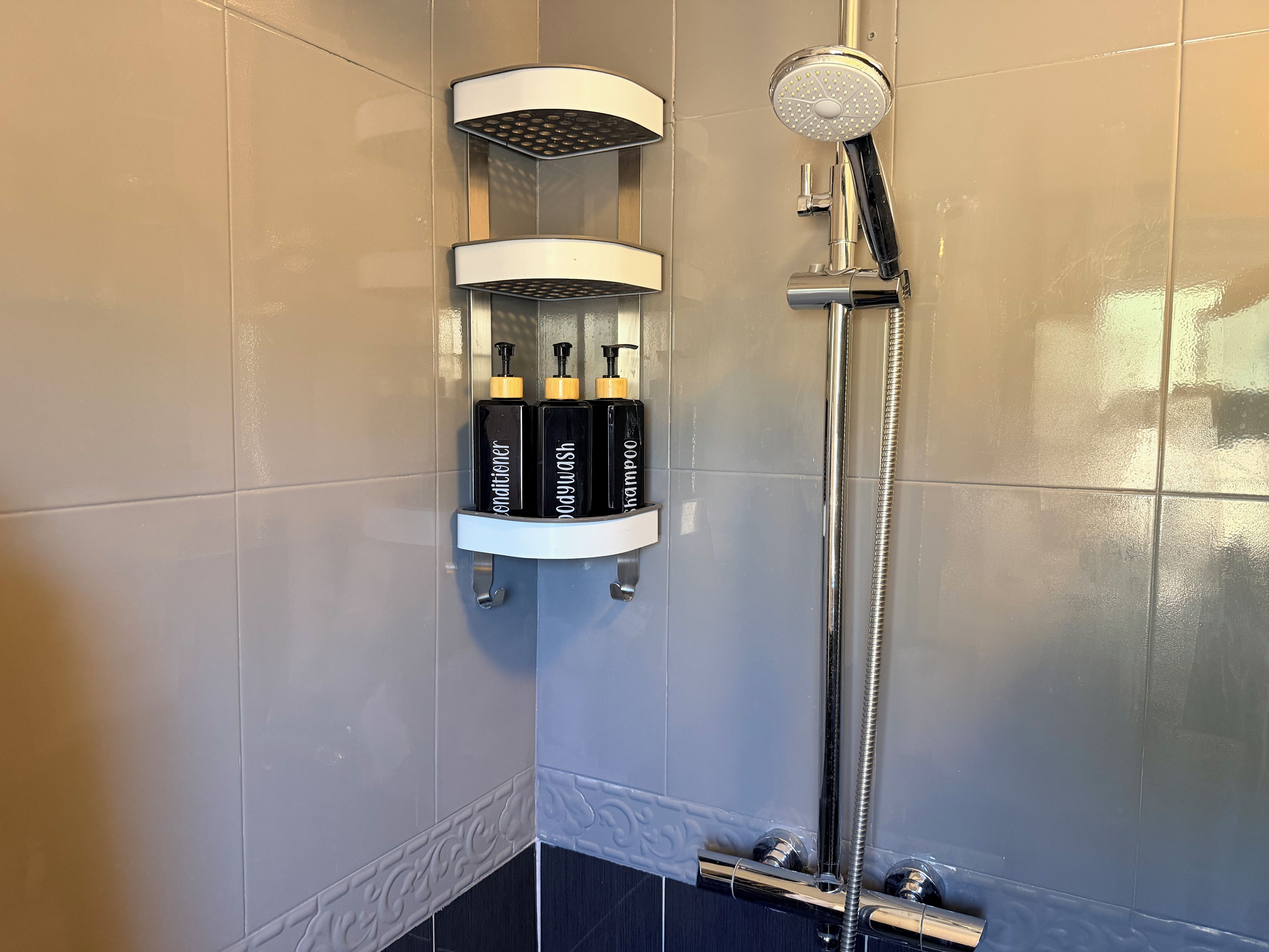 A bathroom with a shower, a white shelf with three bottles, and a tiled wall.