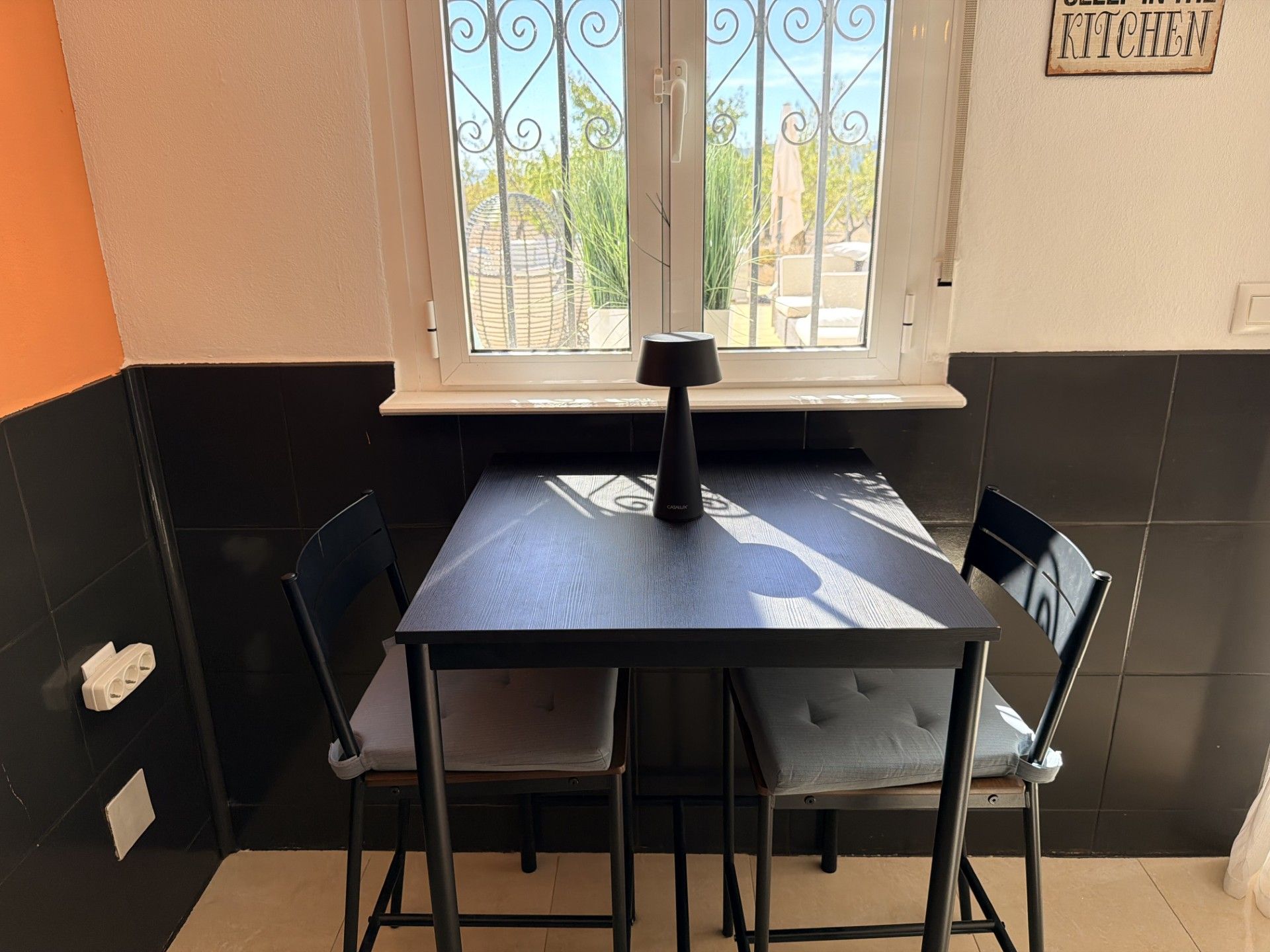 Un pequeño comedor con una mesa y cuatro sillas, un ventanal con vistas a zonas verdes, y un cartel en la pared.