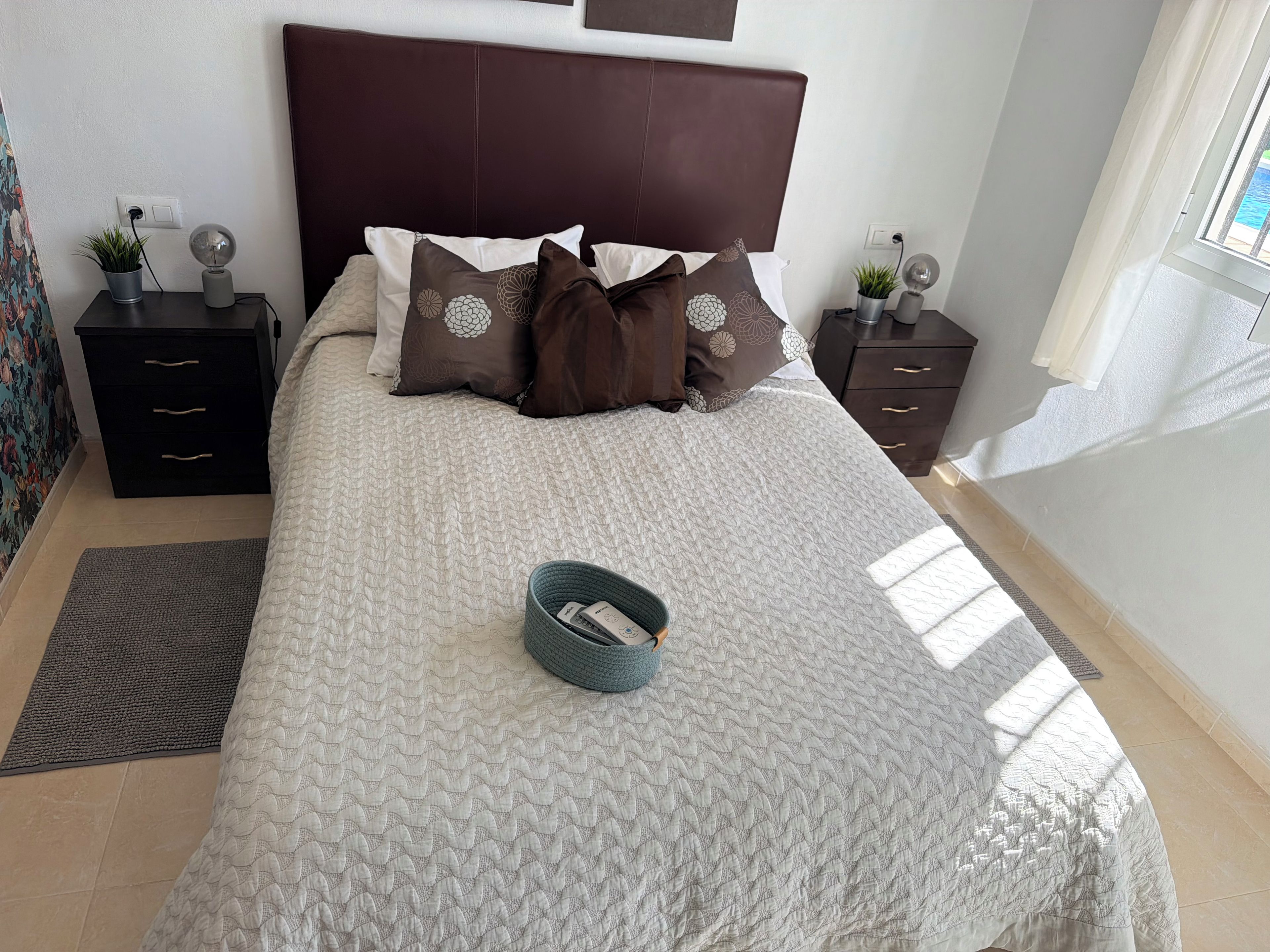 Una cama bien tendida con una colcha estampada, dos mesitas de noche con lámparas, una pequeña planta en maceta y una ventana con vistas.