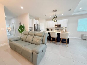 A modern living room with a beige sofa, a kitchen area with white cabinets, and a dining space with two bar stools.
