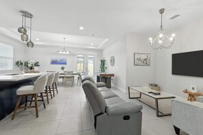 Ein modernes Wohnzimmer mit grauem Sofa, einem Essbereich mit Tisch und Stühlen, einer Küche mit Bar und einem großen Fernseher an der Wand.