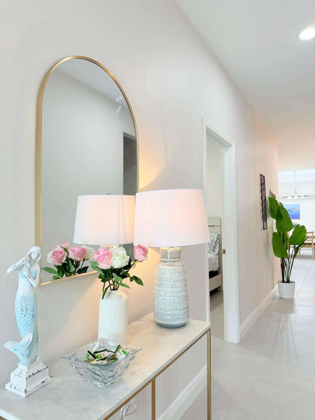 Un couloir avec une console blanche, un grand miroir, deux lampes, un vase de fleurs et une statue décorative représentant une sirène.