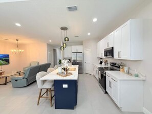 A modern kitchen with white cabinetry, a central island, and a dining area with a grey sofa and a TV.