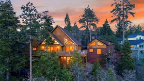 A two-story log cabin with a balcony, surrounded by tall trees and a sunset sky. - Sugar Pine by Avantstay Expansive Deck + Outdoor Dining + Fireplace + Game Room Near Pine Knot Village (Big Bear Lake)