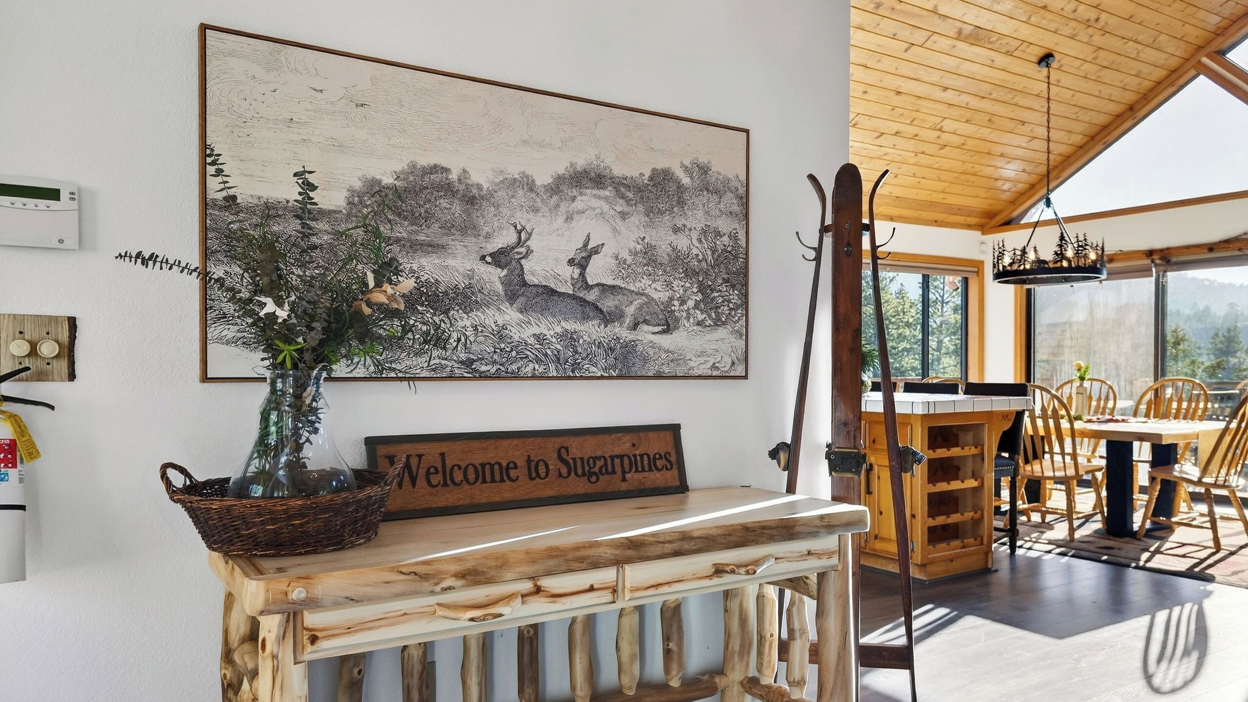A rustic interior with a welcome sign, a wooden table, chairs, and a large landscape painting.