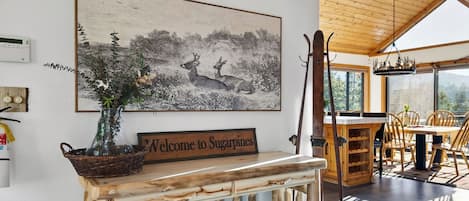 A rustic interior with a welcome sign, a wooden table, chairs, and a large landscape painting.