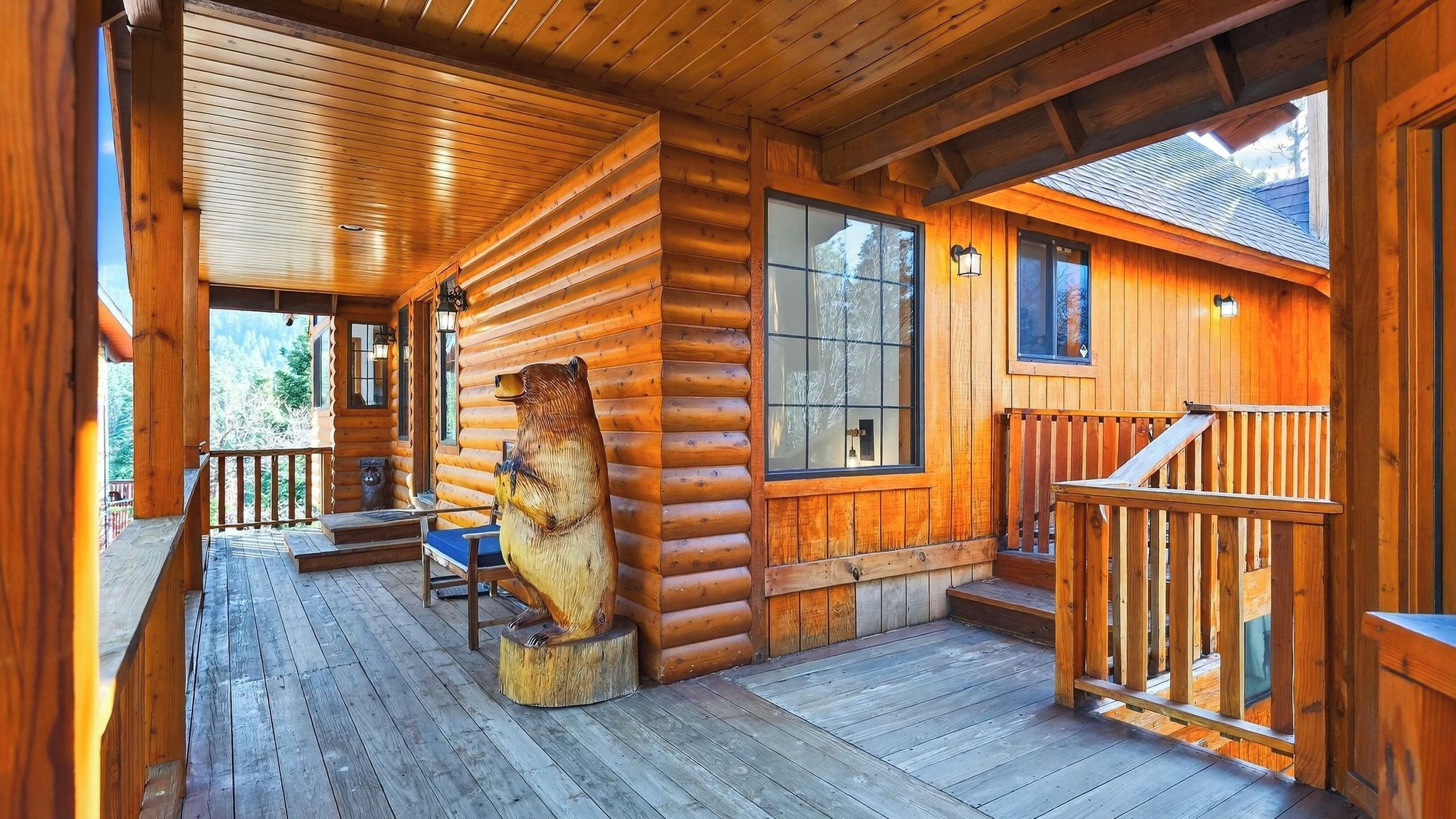 A log cabin with a wooden deck, a large bear sculpture, and a staircase leading to the upper level.
