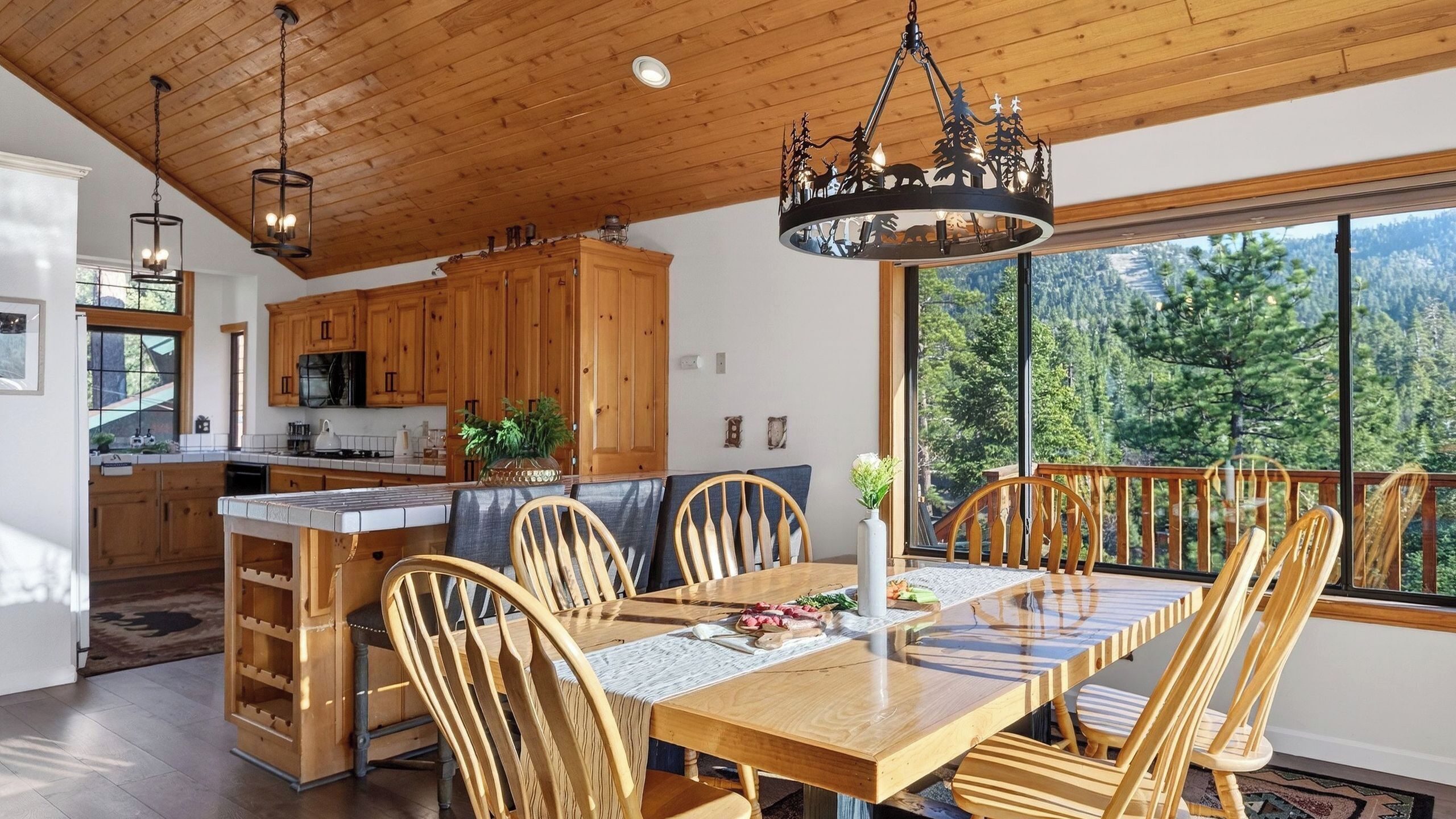 A wooden interior with a dining table, chairs, and a view of a forest through large windows.