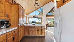 A spacious kitchen with wooden cabinets, a central island, and a large window offering a view of the outdoors. - Sugar Pine by Avantstay Expansive Deck + Outdoor Dining + Fireplace + Game Room Near Pine Knot Village (Big Bear Lake)
