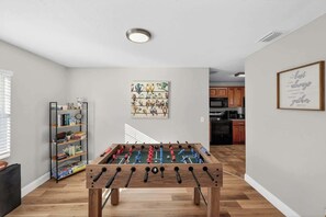 Una habitación con mesa de fútbol, estantería y cocina al fondo.