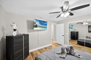 Un dormitorio con cama, cómoda, ventilador de techo, televisión y espejo que refleja la habitación.