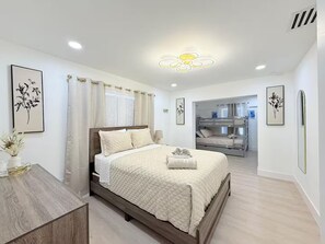Ein Schlafzimmer mit Bett, Nachttisch und Blick in einen Raum mit Etagenbetten.