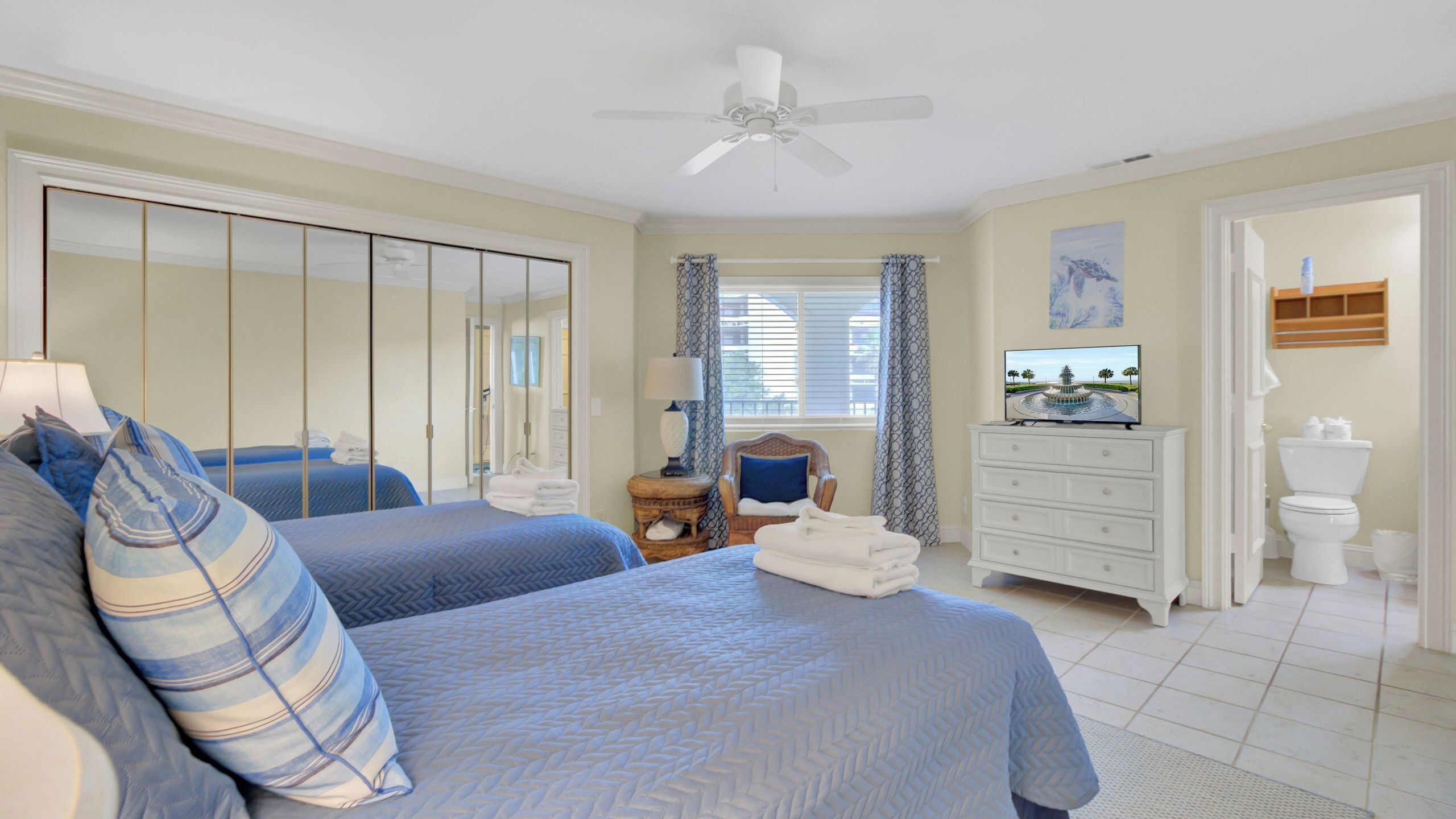 A bedroom with two beds, a ceiling fan, a TV, a dresser, and a bathroom with a toilet and sink.