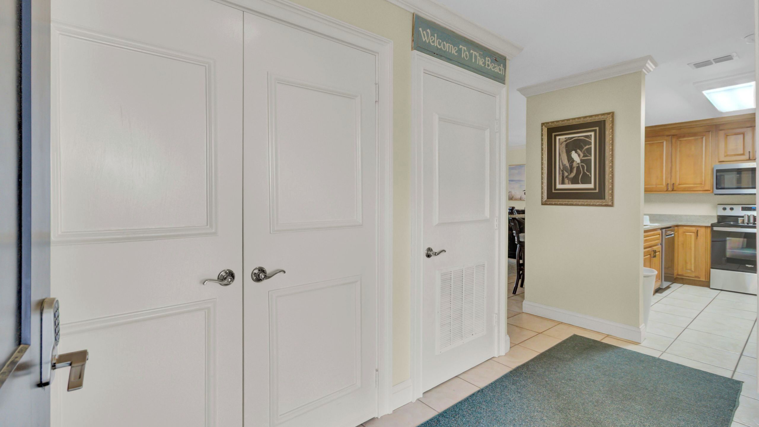 A hallway with double doors, a welcome sign, a framed picture, and a kitchen area with wooden cabinets and appliances.