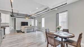 A modern kitchen with white cabinets, a dining area with a round table and chairs, and a TV in the background. - 902 Howard Lofts (Biloxi)