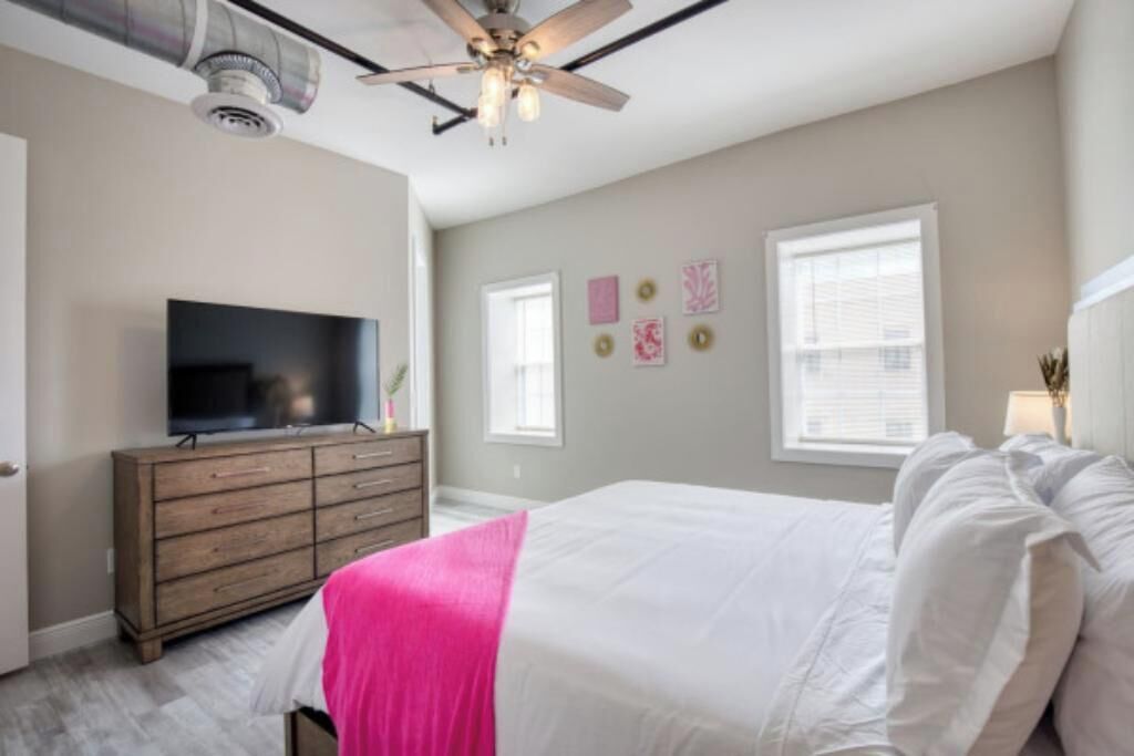A bedroom with a bed, dresser, TV, ceiling fan, and artwork on the wall.