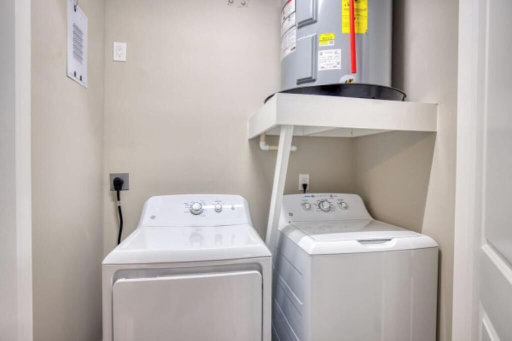 A laundry room with two white washing machines, a water heater, and a white shelf.