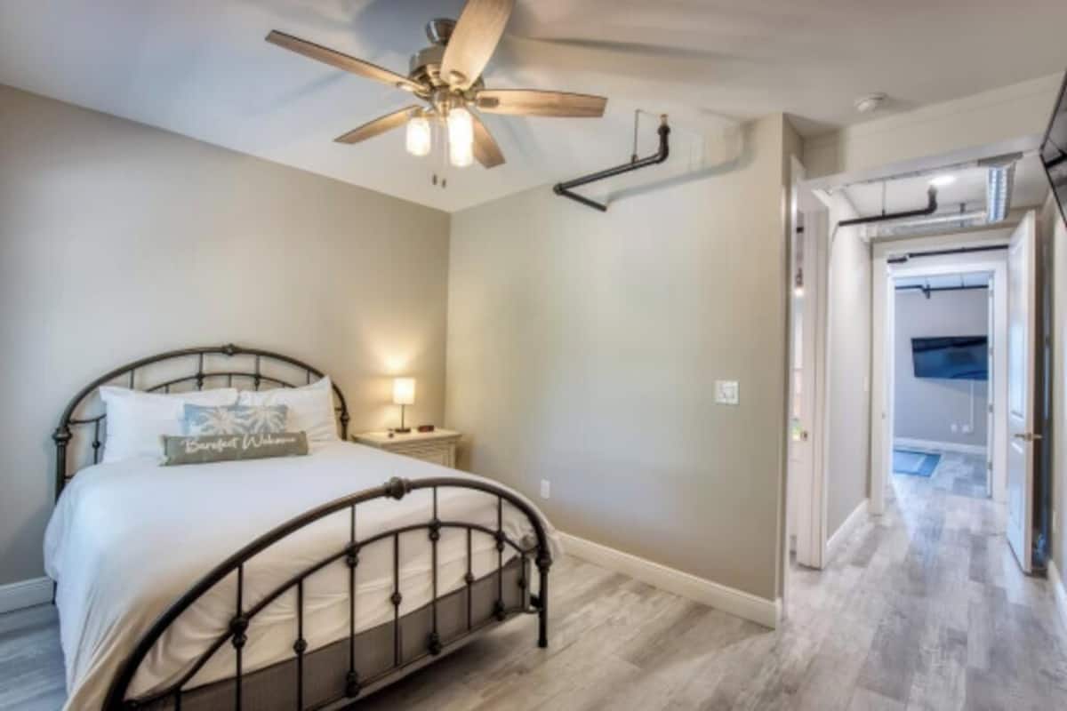 Un dormitorio con cama, ventilador de techo, mesita de noche y una puerta que da a otra habitación.