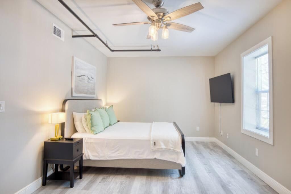 A bedroom with a bed, nightstand, lamp, ceiling fan, artwork, and a TV mounted on the wall.