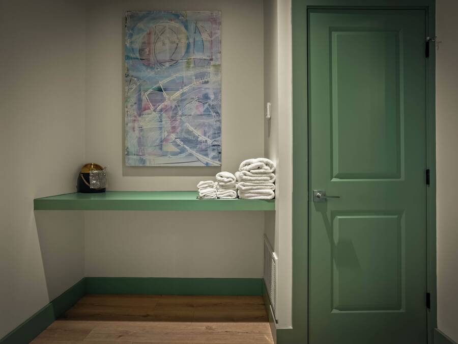 A bathroom with a green door, a towel rack with folded towels, a small shelf with a bag and a container, and a wall-mounted abstract painting.