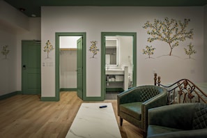 Ein Zimmer mit Holzboden, einem weißen Marmortischchen, grünen Polsterstühlen und einem Bett mit Metallkopfstück.
