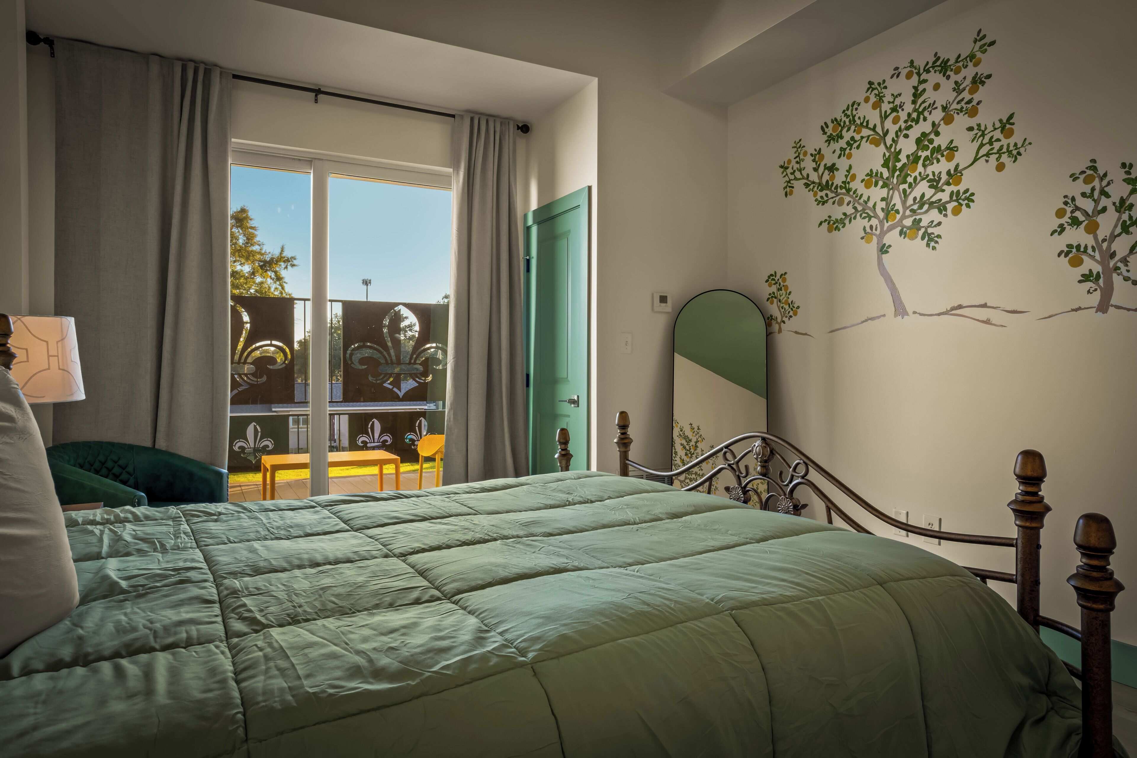 Une chambre avec un lit, une fenêtre avec des rideaux, une porte verte, un mur orné d’une fresque représentant un arbre, et une petite table avec des chaises à l’extérieur.