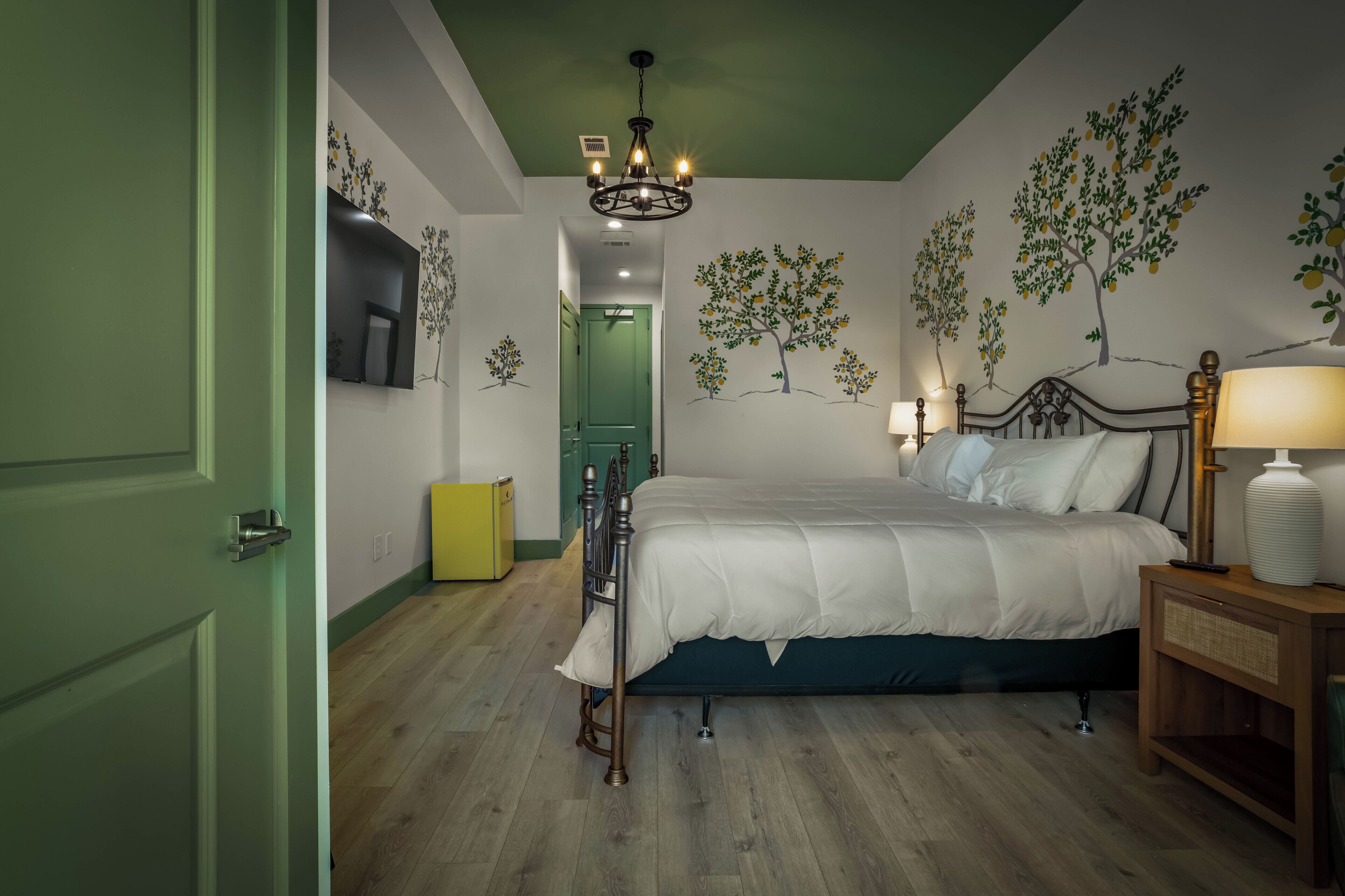 Un dormitorio con cama, lámparas de noche, mesita de noche, una puerta verde y un muro con murales de árboles.
