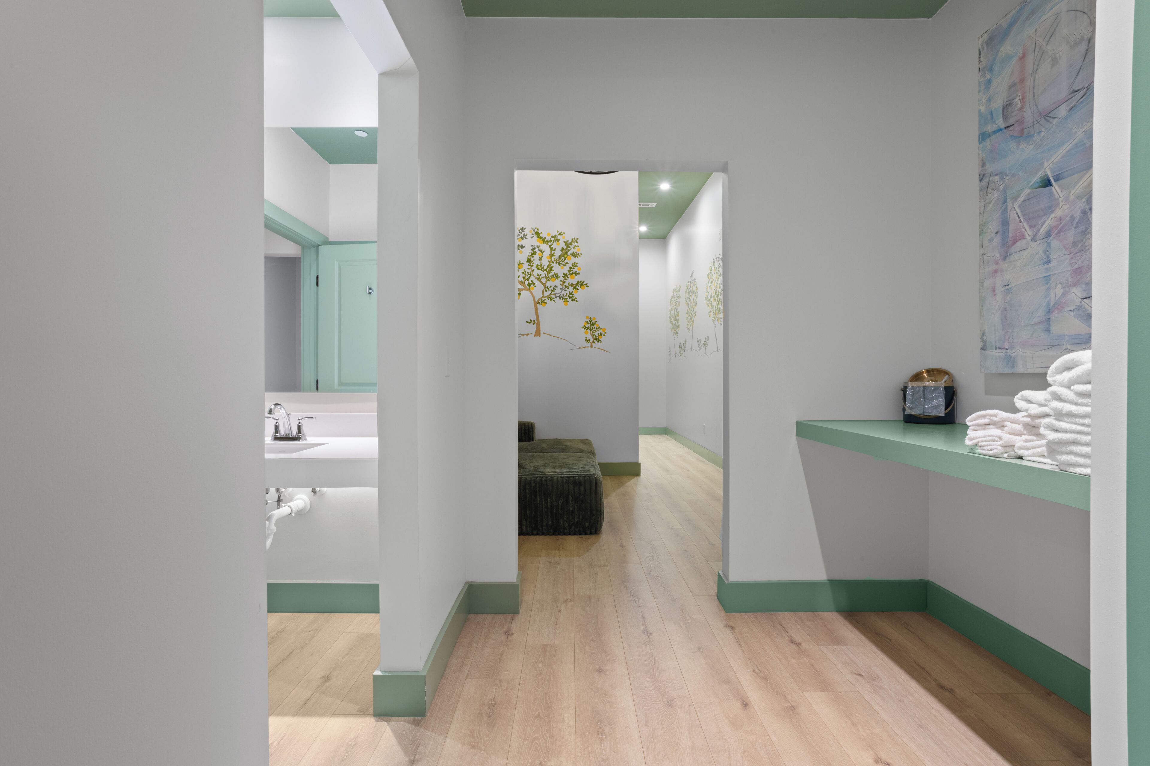 A hallway with a wooden floor, a green-framed mirror, a sink, and a wall with a tree decal.
