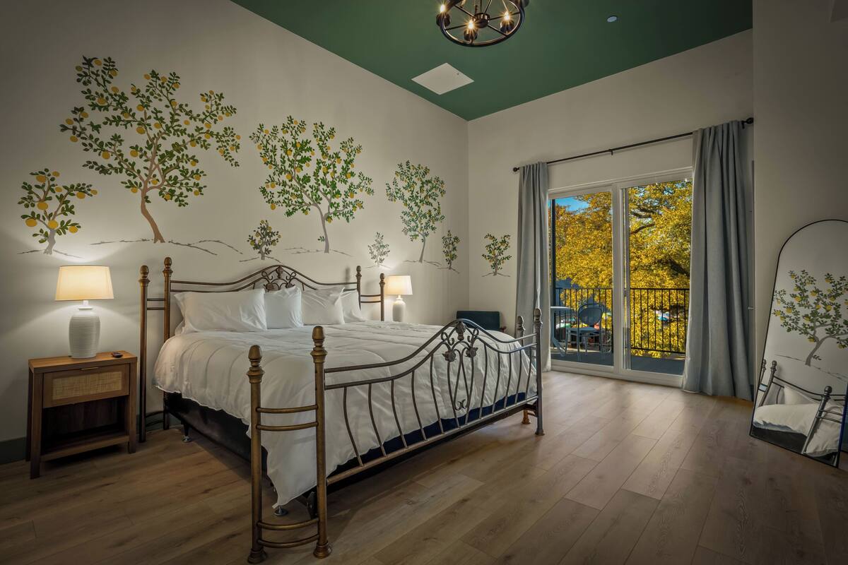 Ein Schlafzimmer mit einem Bett, Nachttischen, einer Chandelier, einem großen Fenster mit Blick und Wandbildern.
