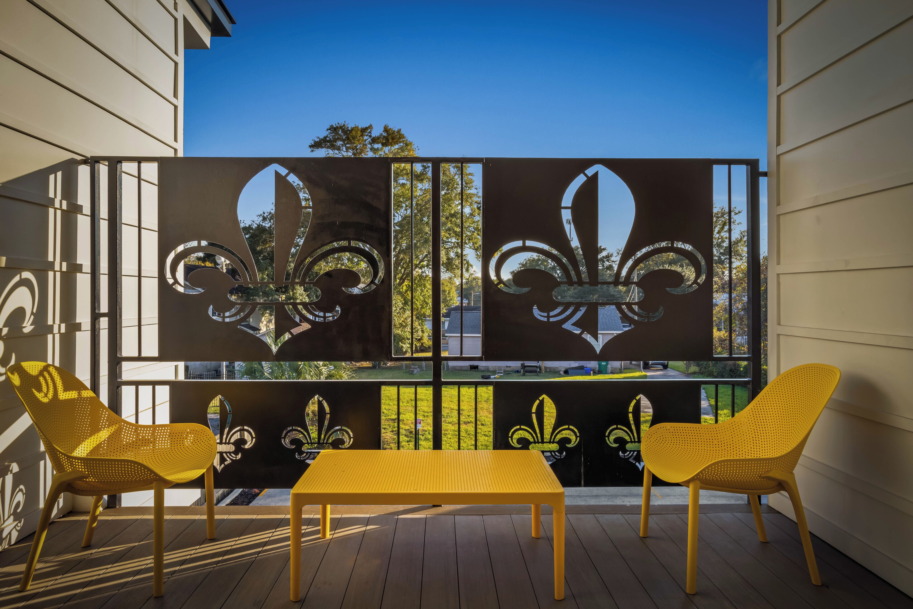 A balcony with a wooden table and two yellow chairs, a decorative metal screen with fleur-de-lis designs, and a view of trees and a building in the background.