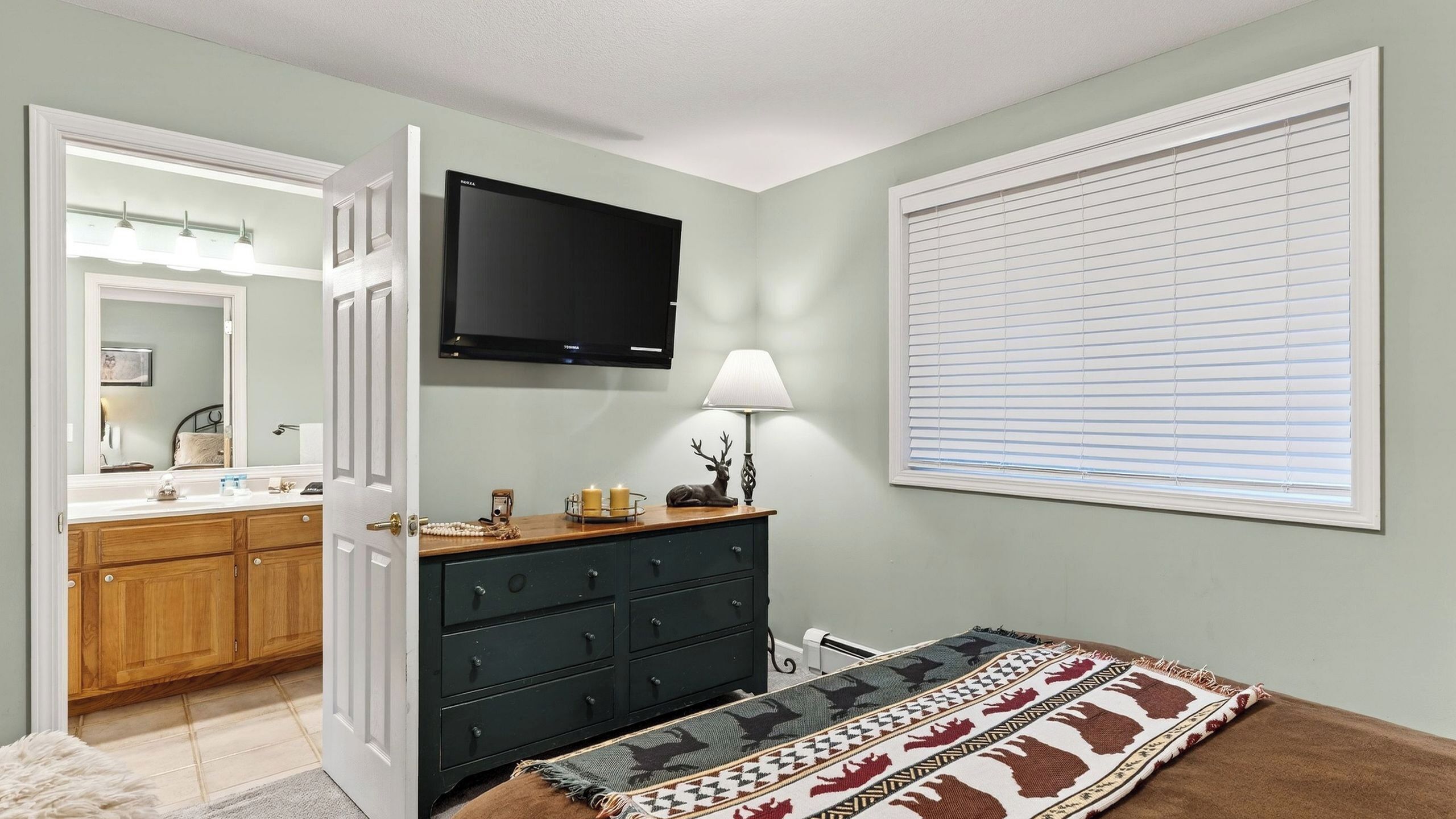 A bedroom with a dresser, a television, a lamp, a window with blinds, and a rug.