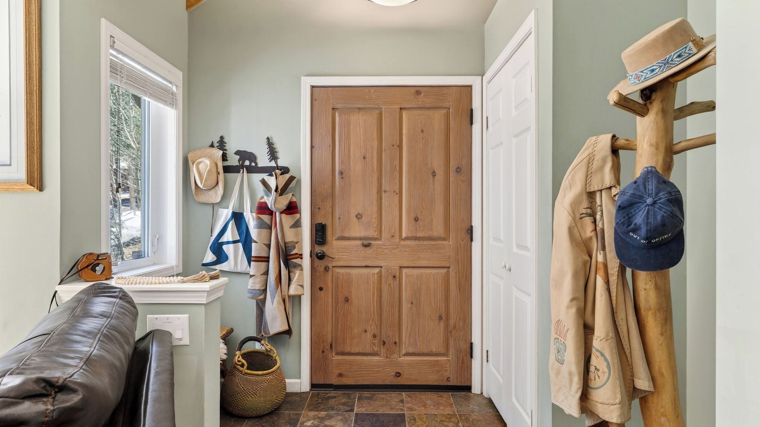 A hallway with a wooden door, a coat rack with hats, a window with blinds, and a leather couch.
