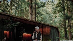 A person standing on a wooden deck in a forested area with a rustic cabin-like structure in the background.