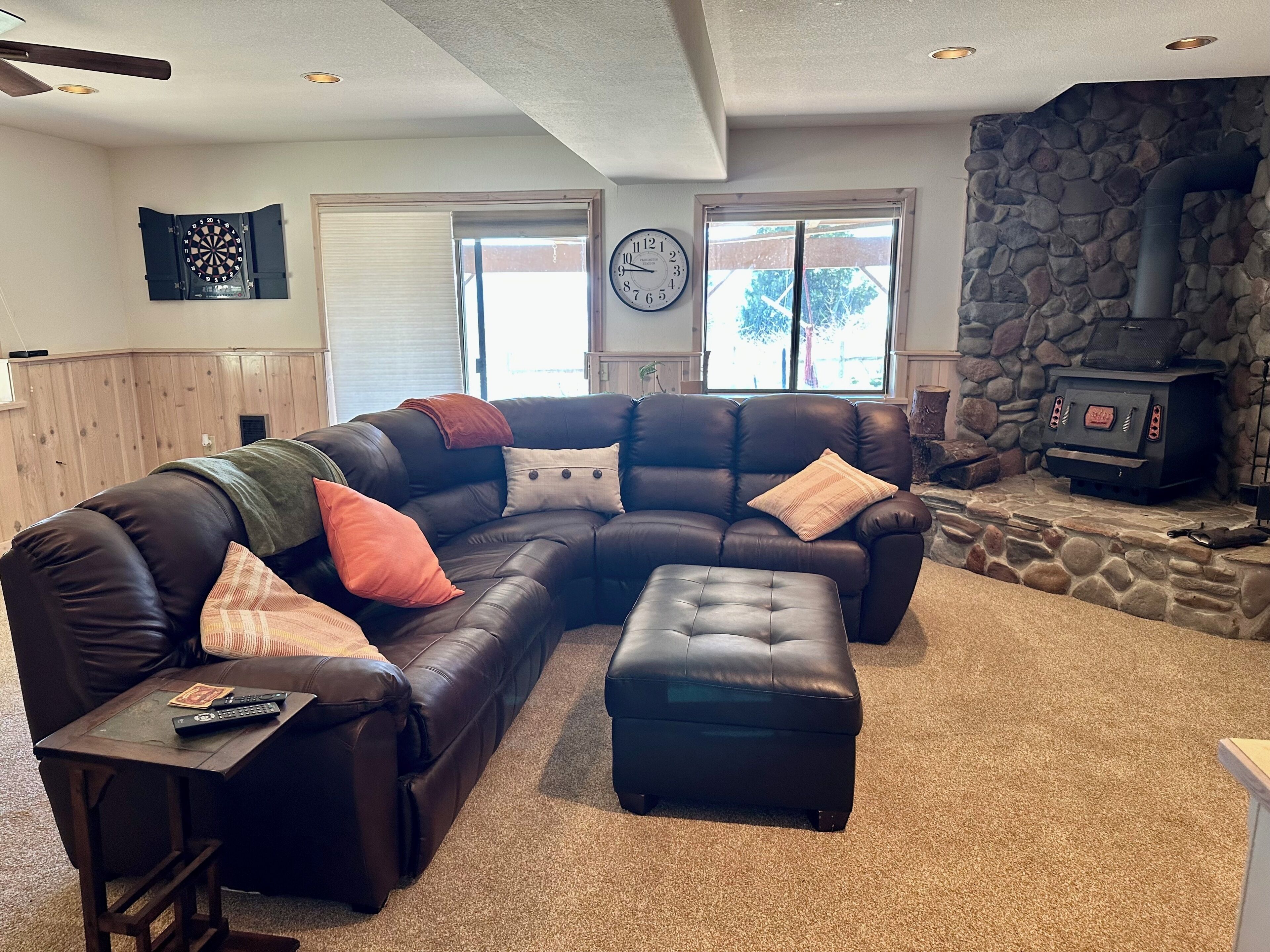 A living room with a sectional sofa, ottoman, stone fireplace, and a dartboard.