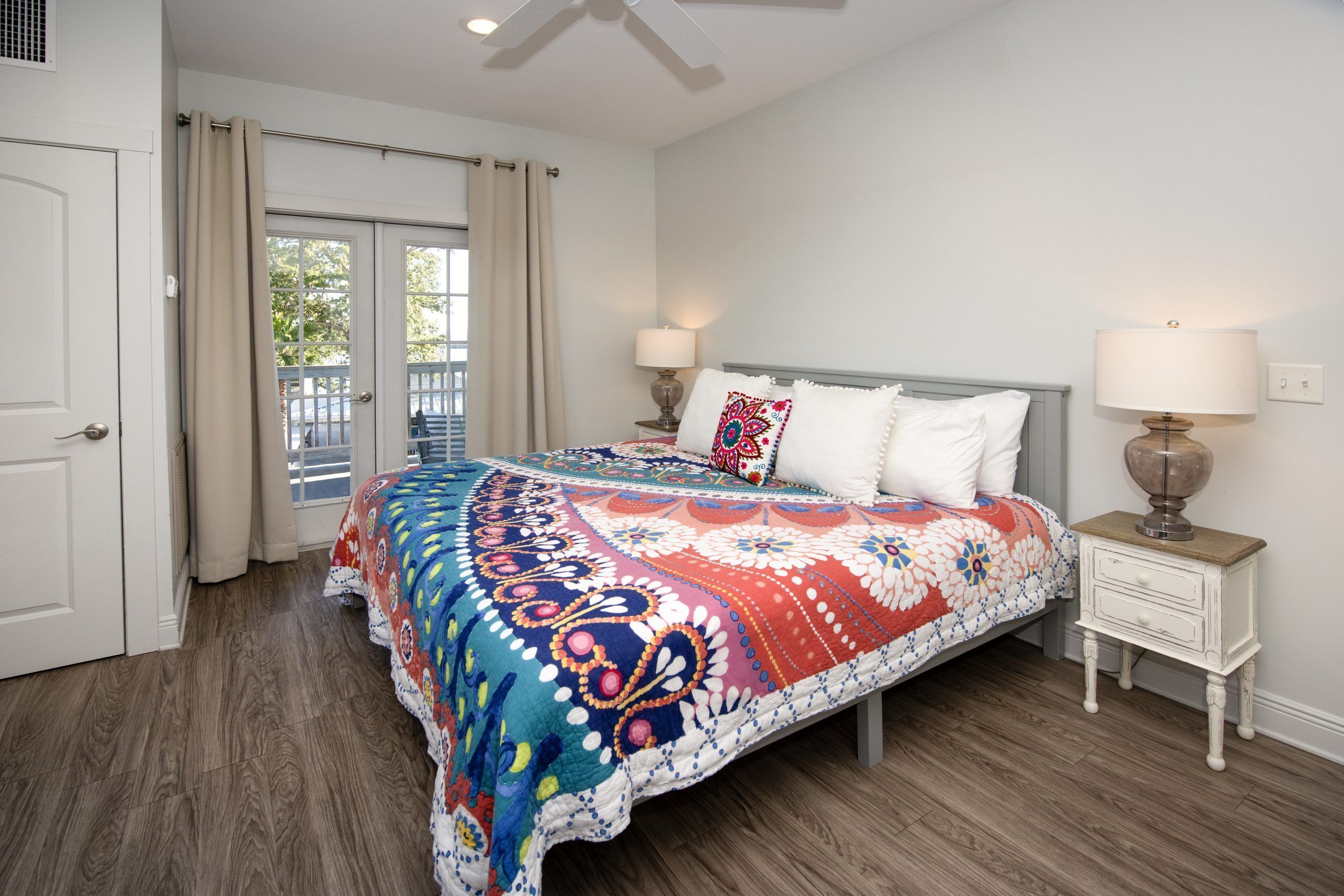 A bedroom with a large bed, two bedside lamps, a nightstand, a door, and a view of the outdoors.