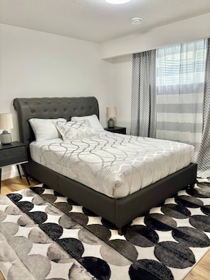 Quarto moderno com cabeceira estofada, colcha estampada, tapete redondo e mesa de cabeceira com abajur.