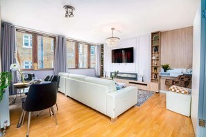 Ein modernes Wohnzimmer mit einer Couch, einem Esstisch, Stühlen, einer Küchenzeile und einem großen Fenster mit Vorhängen.