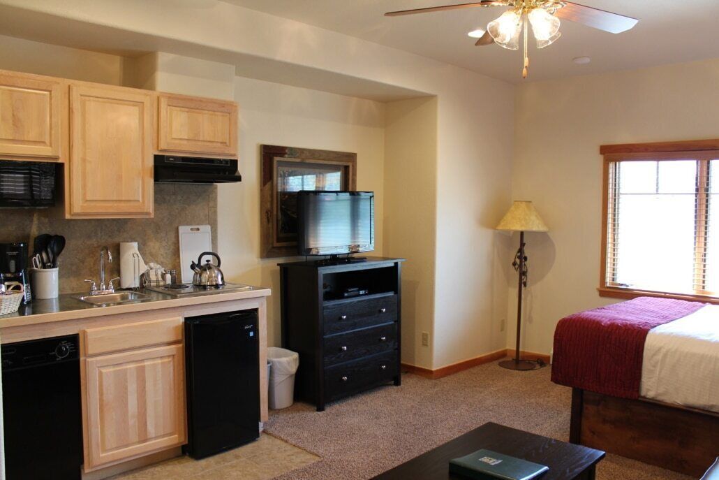 A compact kitchen with wooden cabinets, a dishwasher, a refrigerator, and a television. There is a bed with a red blanket and a lamp near a window.