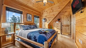 Dormitorio de una cabaña de madera con cama, ventilador de techo, televisor y ventana.