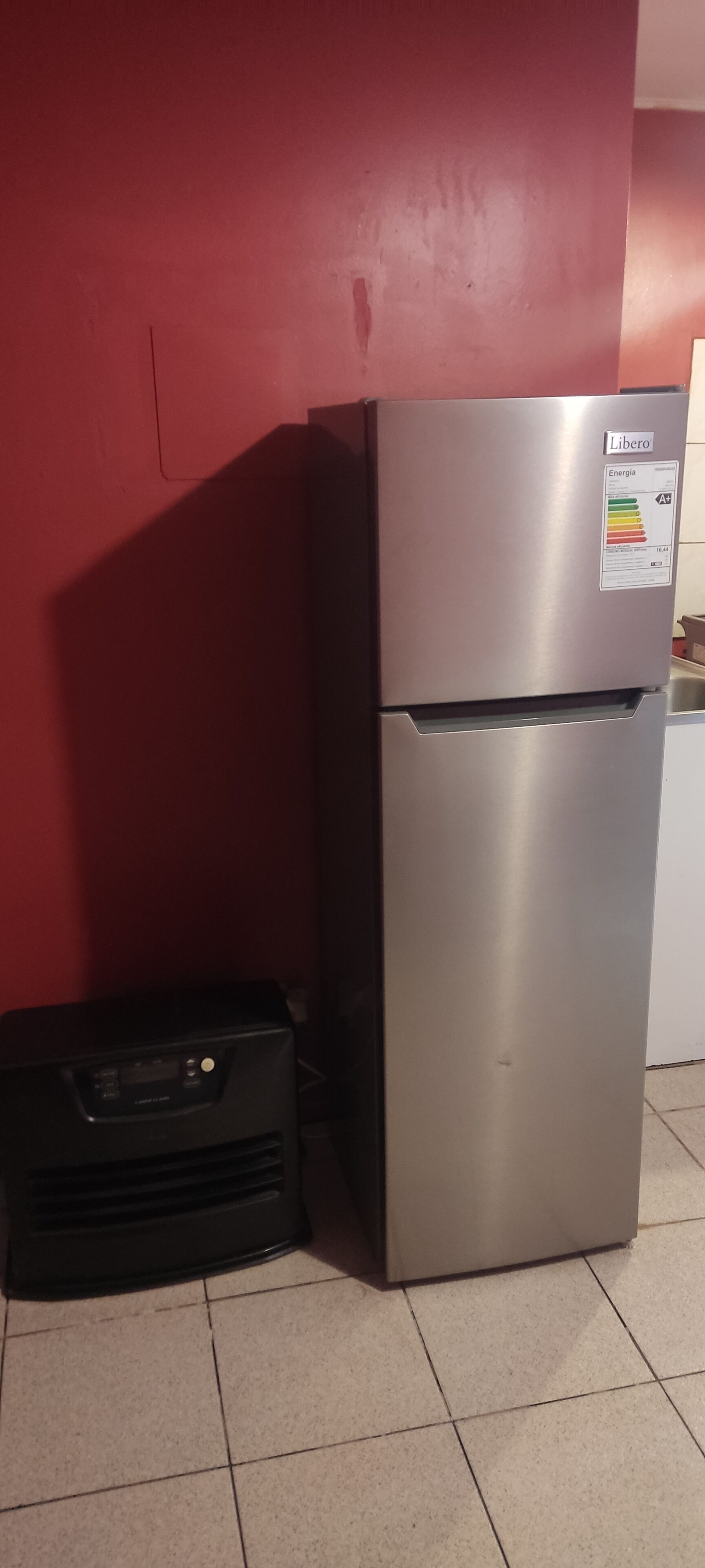 A stainless steel refrigerator with a sticker indicating energy efficiency, a black appliance next to it, and a red wall in the background.