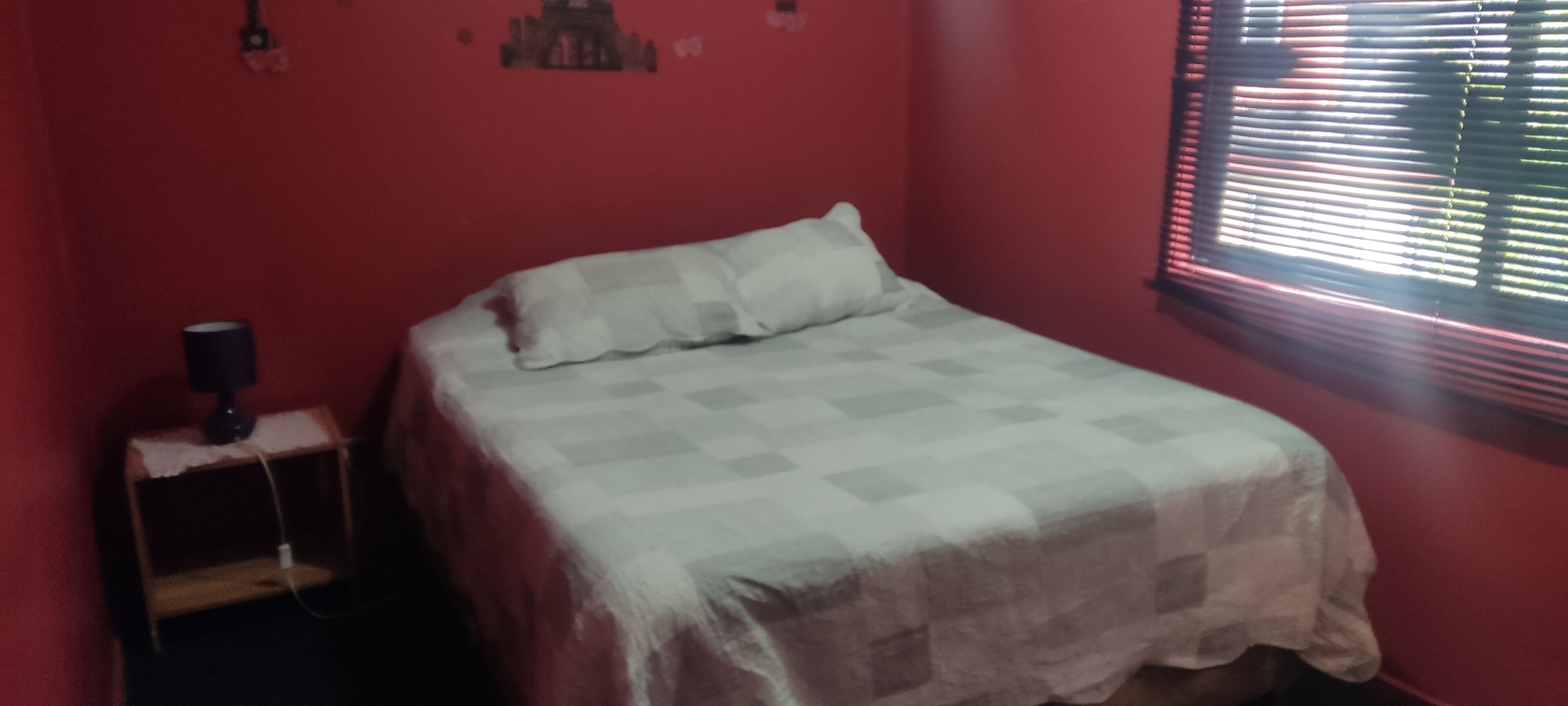 Une chambre avec un lit, une petite table avec une lampe et une fenêtre avec des stores.